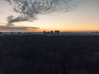 Fototapeta na wymiar Aerial sunset evening view above dark autumn forest near residential Pavlove Pole district in Kharkiv city. Gray multistory buildings with scenic orange cloudy sky on horizon
