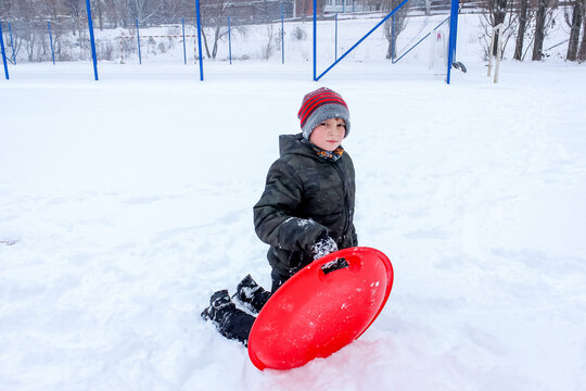 little boy playing in the snow, wearing a red hat, snowy, with a plasma plate for a slide,s kating rink  new year, christmas
