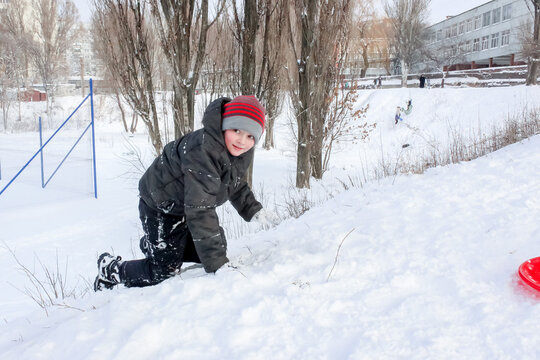 little boy playing in the snow, wearing a red hat, snowy, with a plasma plate for a slide,s kating rink  new year, christmas