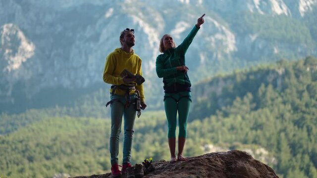two rock climbers stands under rocky cliff, looks at challenging route and tries to read movements and grips. active couple outdoors training on natural rocks with mountain landscape on background