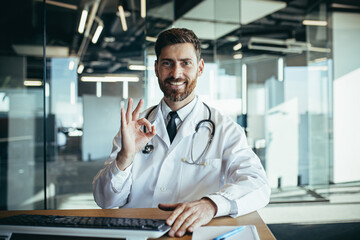 Successful doctor works sitting at the table looks at the camera and smiles makes a positive gesture with his hands happy, works in a modern clinic