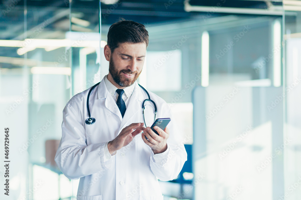 Wall mural male doctor in a white coat in a modern clinic reads information from a mobile phone corresponds wit