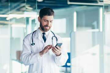 Male doctor in a white coat in a modern clinic reads information from a mobile phone corresponds...