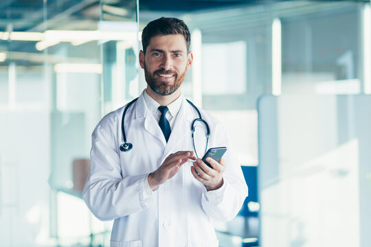 Male doctor in a white coat in a modern clinic reads information from a mobile phone corresponds with colleagues happy and smiles - Powered by Adobe