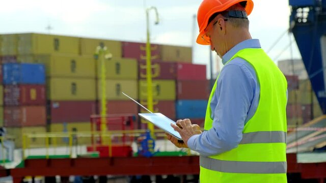 Marine port cargo industry. Mature employee in protective helmet with tablet computer counts large metal containers in modern dockyard