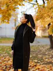 Young beautiful pregnant woman with dark hair in a black tight dress and coat posing on an autumn meadow in the park