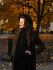 Young beautiful pregnant woman in a hat with dark hair in a black tight dress and coat posing on an autumn meadow in the park