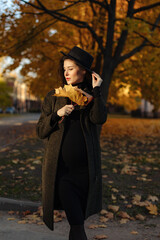 Young beautiful pregnant woman in a hat with dark hair in a black tight dress and coat posing on an autumn meadow in the park