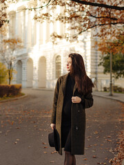 Fototapeta premium Young beautiful pregnant woman with dark hair in a black tight dress and coat posing on an autumn meadow in the park
