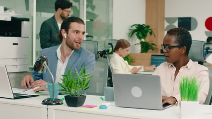 A very handsome man with brown hair tied back and a beard is in a very large aesthetic office, he is talking with his co-worker who is a very beautiful woman wearing glasses