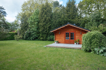 Orange wooden hut in the garden with many tall trees. Garden shed with lawn in front of him	
