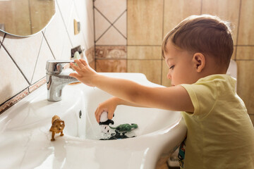 Boy in yellow t-shirt washing animal plastic toys in white sink 