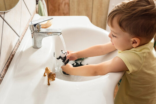 Boy in yellow t-shirt washing animal plastic toys in white sink 