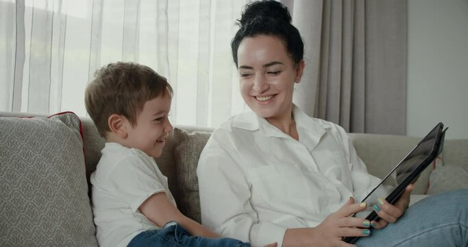 Happy mom and cute son little child, mother plays with her baby at home, relaxing, using smartphone, hugging, sit on the sofa, boy, watch funny videos, have fun, enjoy the moments of family life.