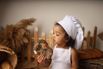 a cute dark-skinned baby with curly hair in a chef's costume has a bagel and a roll. High-quality...