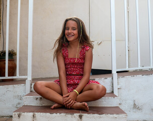 A low angle shot of a young Caucasian girl wearing a red sundress and barefoot, sitting on the stairs and smiling at the camera