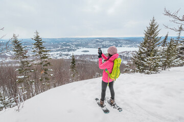 Phone. Hiking snowshoeing woman in winter forest using phone app taking photo of idyllic winter landscape. People on hike in snow living active lifestyle. Mont Tremblant, Laurentians, Quebec, Canada