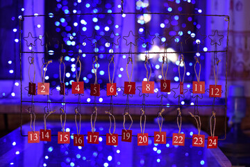 Christmas Advent calendar with numbers on red plaques hangs against blurred window with blue garlands. Family tradition.