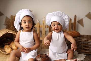 two cute dark-skinned kids with curly hair in a cook's costume are eating a bagel and a roll. High-quality photography