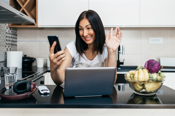 Beautiful middle age woman sitting alone in her apartment and enjoying in free time. She using laptop computer and tablet for chat and music listening.