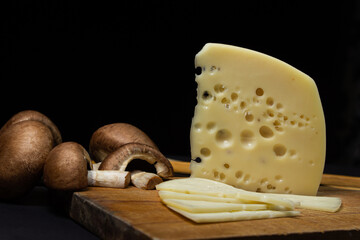 Yellow cheese on a black background. Sliced cheese next to raw mushrooms on a dark background. Cheese with ingredients on wooden board