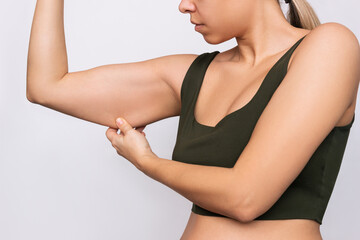 Fototapeta na wymiar Cropped shot of a young caucasian blonde woman grabbing skin on her upper arm with excess fat isolated on a gray background. Pinching the loose and saggy muscles. Taking care of the body