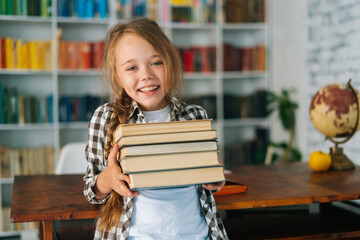 Medium shot portrait of joyful elementary child school girl holding stack of books in library at school, looking at camera. Cute primary pupil schoolgirl standing on blurry background of bookshelf.