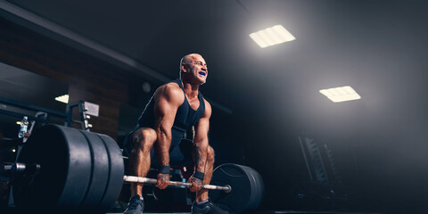 Fototapeta na wymiar Emotional Older sportsman exercising deadlift with barbell while on cross training in a gym. 