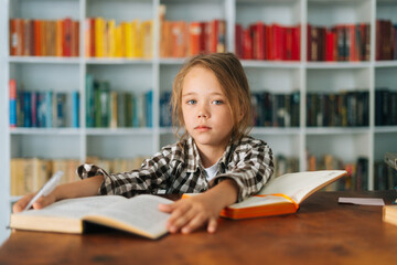 Front view of pretty pupil school girl kid doing homework reading paper book sitting at table in light children room. Portrait of elementary child schoolgirl studying at home, selective focus - obrazy, fototapety, plakaty