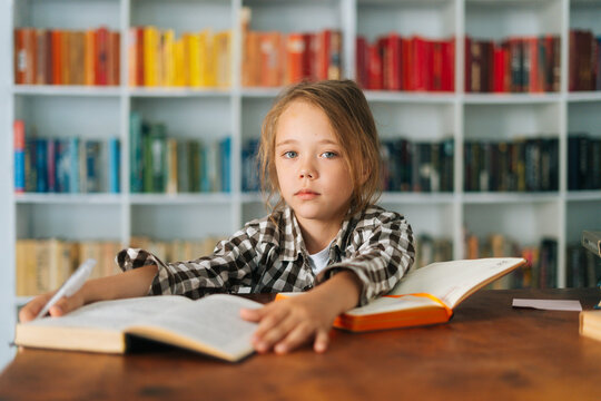 Front view of pretty pupil school girl kid doing homework reading paper book sitting at table in light children room. Portrait of elementary child schoolgirl studying at home, selective focus