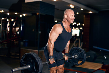 Fototapeta na wymiar Adult bald Bodybuilder muscular man doing heavy deadlift exercise with weight while in gym in dark 
