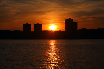 Fototapeta premium The orange and round sun going behind the skyscrapers and illuminating the river