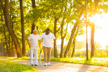 Caregiver is teaching old man to walk with walker. Professional nurse and patient walking outdoor at sunset. Assistance, rehabilitation and health care.