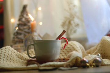 Obraz na płótnie Canvas Cup of warm drink with candy cane and cinnamon stick, soft blanket and various Christmas decorations. Hygge at home. Selective focus.