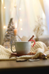Cup of warm drink with candy cane and cinnamon stick, soft blanket and various Christmas decorations. Hygge at home. Selective focus.