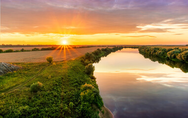 Scenic view at beautiful summer river sunset with reflection on water with green bushes, grass, golden sun rays, calm water ,deep cloudy sky and glow on a background, spring , evening landscape