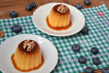 Flan with a walnut on top on a green and white checkered tablecloth surrounded by blueberries