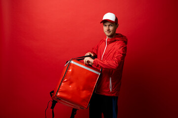 Delivery employee man 20s in red uniform with a thermal food bag backpack works in a courier service isolated on red background studio.