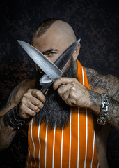 A brutal sexy man with a black beard in an apron has crossed big knives and looks menacing