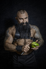 A brutal sexy man with a black beard with a naked hairy torso cuts a green apple with a large knife