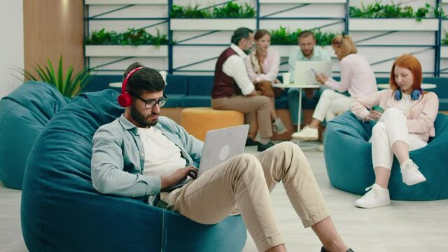A very smart looking brunette man with a beard and glasses is doing work seriously on his laptop while listening to music on his headphone and sitting on a bean bag chair. Arri Alexa Mini.