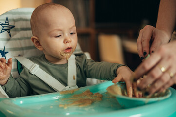 Baby eating with a face stained in food while mother feeds him with a spoon