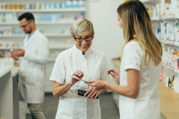 Senior female pharmacist using a tablet while working in a pharmacy with her young colleague