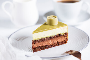 Pistachio chocolate cake with four layers and green pistachio glazing on top served on a white plate on white background with coffee in blur