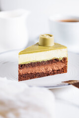 Pistachio chocolate cake with four layers and green pistachio glazing on top served on a white plate on white background with coffee in blur
