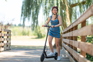 A girl in a safety helmet rides an electric scooter near a large wooden fence. Beautiful nature. Leisure activities, sports, vacations, environmentally friendly modern transport.