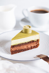 Pistachio chocolate cake with four layers and green pistachio glazing on top served on a white plate on white background with coffee in blur