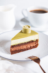 Pistachio chocolate cake with four layers and green pistachio glazing on top served on a white plate on white background with coffee in blur