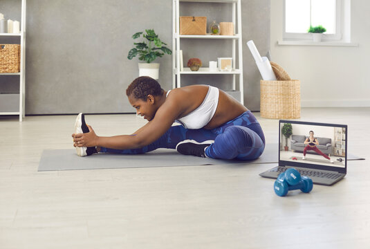 Enduring African American woman who wants to stay fit is training at home using online workout broadcast. Young woman near laptop on sports mat stretches before start of her morning workout.