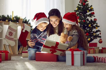 Happy loving mother and small children open gifts near Christmas tree on winter holiday. Smiling mom and little kids unbox presents from Santa Claus on New Year eve. Family celebration concept.